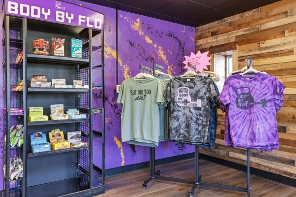 BodyByFlo pro shop with supplement shelving and branded tie-dye t-shirts on racks